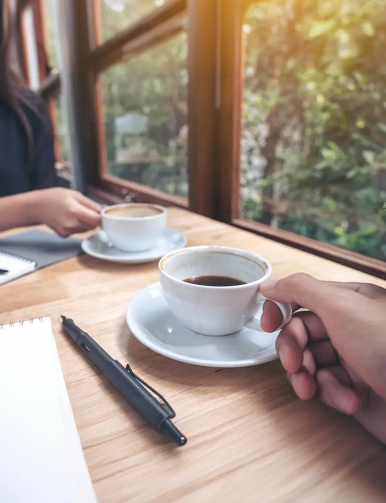 Two people drinking coffee, only hands visible. Both have notepads and pens out and are sat at a wooden table next to a window with lots of green foliage and warm sunlight coming through the window.
